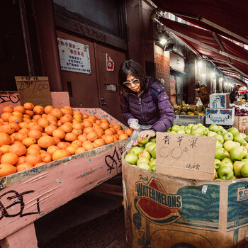 Vendor at a market stall wearing a purple jacket, standing beside wooden crates of oranges and green apples.