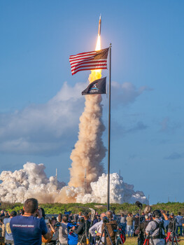 Rocket launch with American flag and military pennant in foreground, spectators watching from ground level.