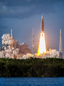 Rocket launching at dawn with bright orange flame and white smoke clouds against blue sky over water.
