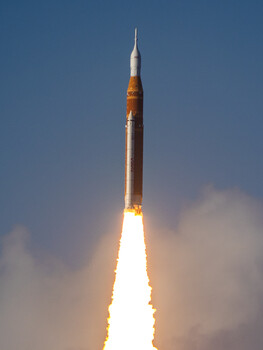 Rocket launching vertically against a blue sky with bright white exhaust flames and orange heat damage visible on the booster.