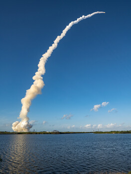 Space shuttle launch creating a towering plume of white smoke and exhaust against a clear blue sky over water.