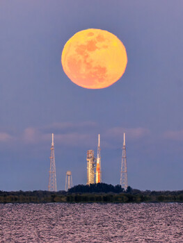 Massive full moon rising above illuminated rocket launch complex with multiple tall towers reflected in water.