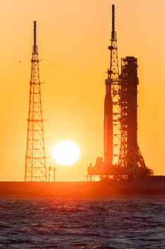 Space launch vehicle and communication tower silhouetted against golden sunset over water.
