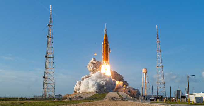 Rocket launching from a coastal pad with massive white smoke plume against clear blue sky and launch towers flanking the site.
