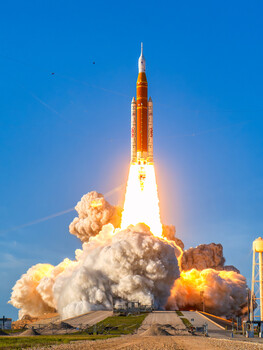 Rocket launching with massive plumes of white and orange smoke against clear blue sky.