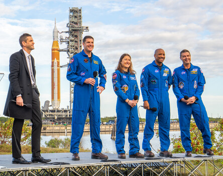 Five people in blue NASA spacesuits stand in front of a tall orange and white launch vehicle at a spaceport.