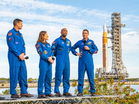 Four astronauts in blue spacesuits standing in front of a tall rocket on launch pad.