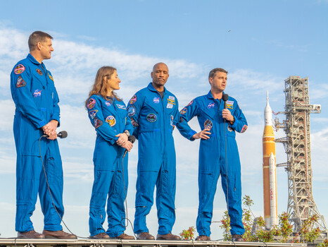 Four astronauts in blue flight suits stand in front of a launch tower under clear skies.
