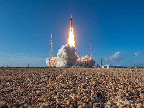Rocket launching from a desert landscape with bright flame and smoke plume against clear blue sky.