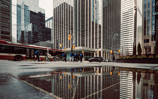 Urban street scene with tall buildings reflected in a wet pavement puddle.