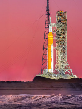 Illuminated rocket on launch pad silhouetted against dramatic pink and purple twilight sky.