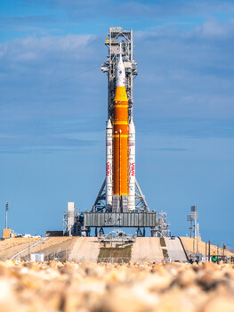 Tall orange and white rocket on launch pad under blue sky with wispy clouds.