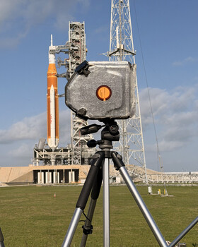 Survey-grade laser theodolite mounted on tripod with rocket and launch tower in background.