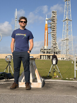 Man in NASA shirt standing on concrete pad with surveying equipment and tall rockets visible in background.