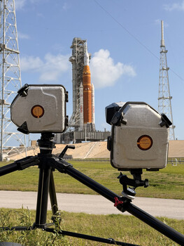 Two thermal imaging cameras mounted on a horizontal surveying pole with launch pad and communication towers visible in the background.