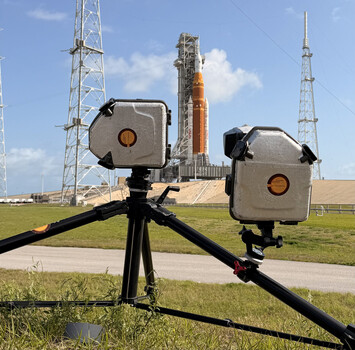 Two thermal imaging cameras mounted on a tripod aimed at a rocket on the launch pad.
