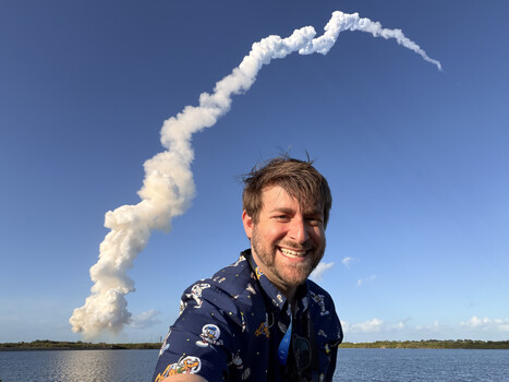 Man in navy patterned shirt smiling at camera with rocket launch plume in background.
