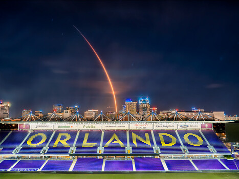 Lightning strike illuminates a stadium at dusk with purple seating and city skyline beyond.