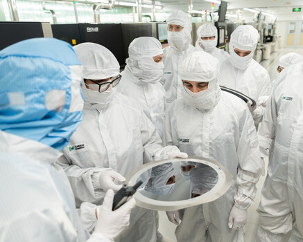 Team of scientists in full protective suits examining a circular wafer or semiconductor during cleanroom inspection.