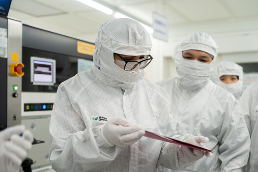 Three technicians in full cleanroom protective gear examine a sample in a laboratory setting.
