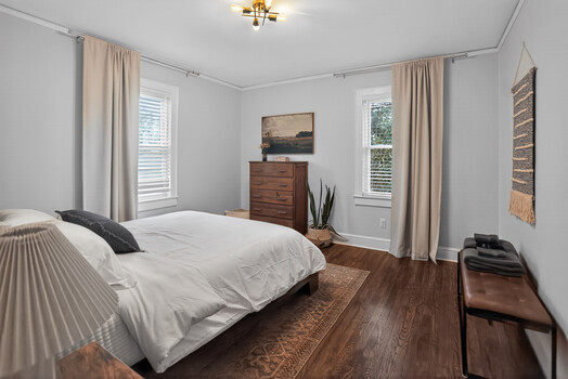Bright, airy bedroom with white walls, wooden dresser, and hardwood flooring bathed in natural light from multiple windows.
