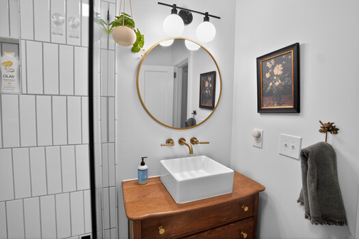 Modern bathroom vanity with wooden base, white vessel sink, brass fixtures, and round mirror with plant.