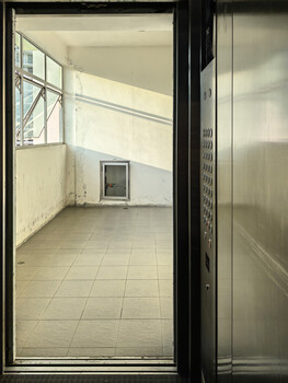 View through a dark-framed glass door into a sparse, minimalist interior room with pale walls and tiled flooring.