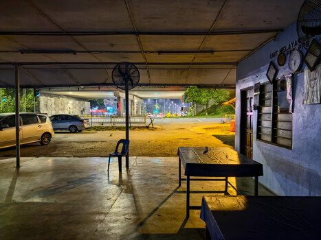 Interior of a covered arena or stable structure with concrete flooring, a black fan, and a wooden bench in the foreground.