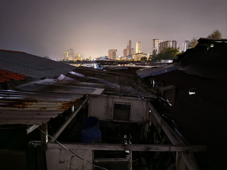 Urban waterfront with dilapidated structures in foreground and illuminated city skyline at dusk.