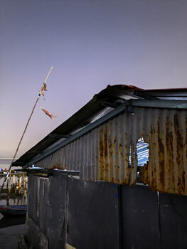 Weathered black industrial structure with antenna against twilight sky and distant harbor lights.