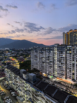 Urban cityscape at dusk with illuminated high-rise residential buildings overlooking tree-covered hills under a purple twilight sky.