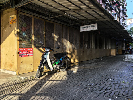 Blue and white scooter parked against a yellow wall with dark storefront shutters, creating strong shadow patterns on brick pavement.