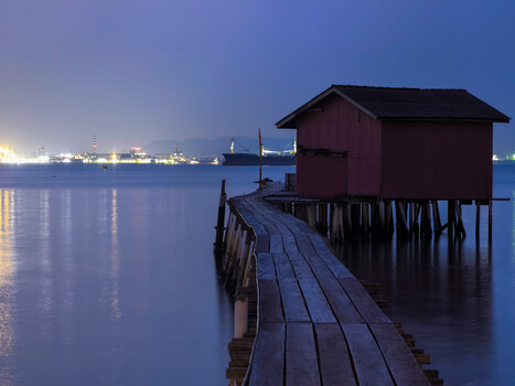 Wooden boardwalk extending toward boathouses at dusk with city lights reflecting on calm water.