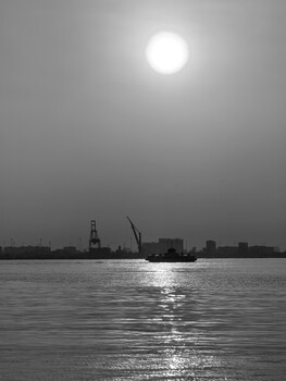 Industrial waterfront at dusk with sun reflecting on calm water and silhouetted cranes and buildings.
