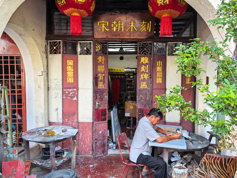 Man working at a table inside a traditional Chinese storefront with red pillars and hanging lanterns.