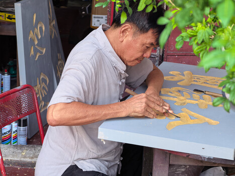 Artisan hand-painting gold calligraphy characters on a white wooden surface outdoors.