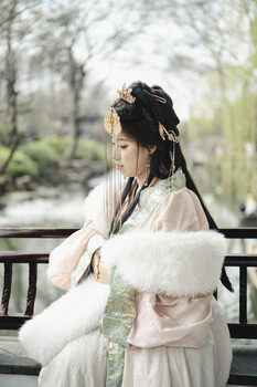Woman in flowing white garments seated on a wooden bench, looking to the side with hair styled in braids.