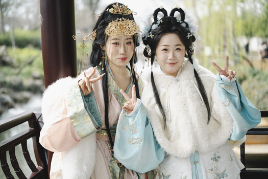 Two women in traditional Chinese costume pose together outdoors, wearing flowing robes with fur stoles and ornate hair accessories.