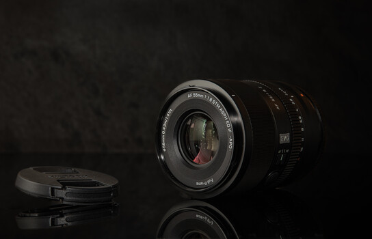 Vintage manual camera lens with lens cap displayed on reflective black surface against dark background.