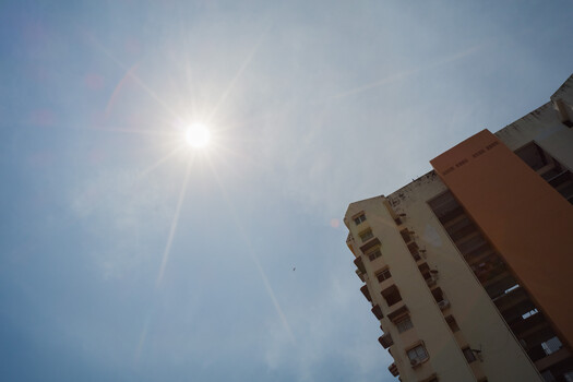 Bright sun blazing in a clear sky above a tan residential building with multiple windows.
