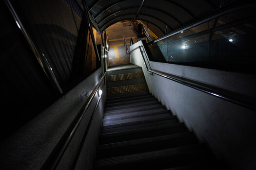 Dimly lit stairwell with metal railings descending into darkness, lit by overhead fixtures.