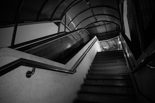 Black and white photograph of a stairwell descending through an industrial tunnel with curved ceiling and metal railings.