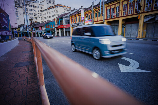 Blue van in motion on urban street with colorful storefronts and red curb in foreground.