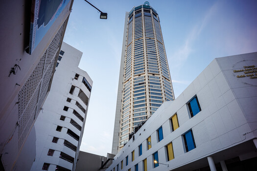 Tall residential tower rising above lower-rise apartment buildings, shot from ground level looking upward against blue sky.