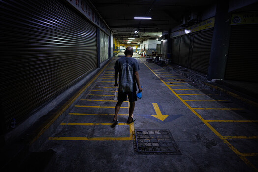 Person walking through an empty covered market corridor with metal shutters and yellow floor markings at night.
