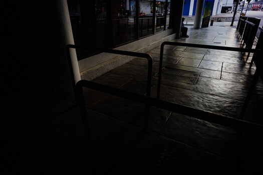 Wet urban pavement at night with metal railings and storefronts in soft background light.