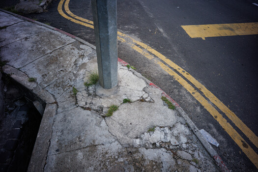 Cracked concrete sidewalk with weeds and a metal utility pole casting shadow, adjacent to yellow no-parking lines on dark asphalt.