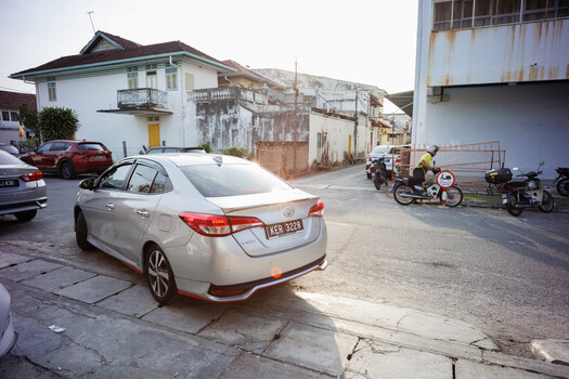 White sedan parked in a deteriorating urban courtyard with decaying buildings and motorcycles in the background.