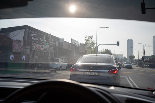 View through a car windshield of urban traffic with a sedan ahead and city buildings in overcast daylight.