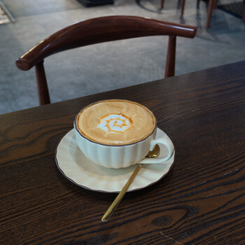 Cappuccino with latte art in a white cup on a wooden table with a wooden chair in the background.